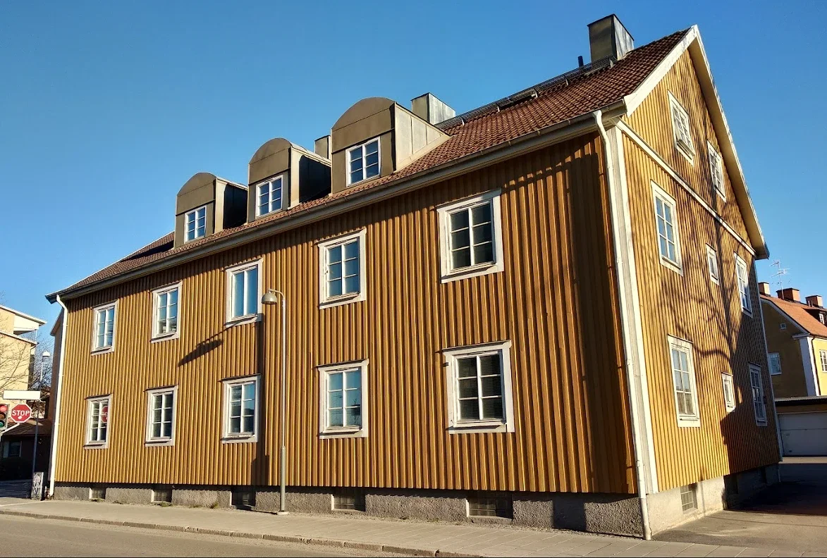 Gul träbyggnad med vertikala paneler och vita fönster under blå himmel.